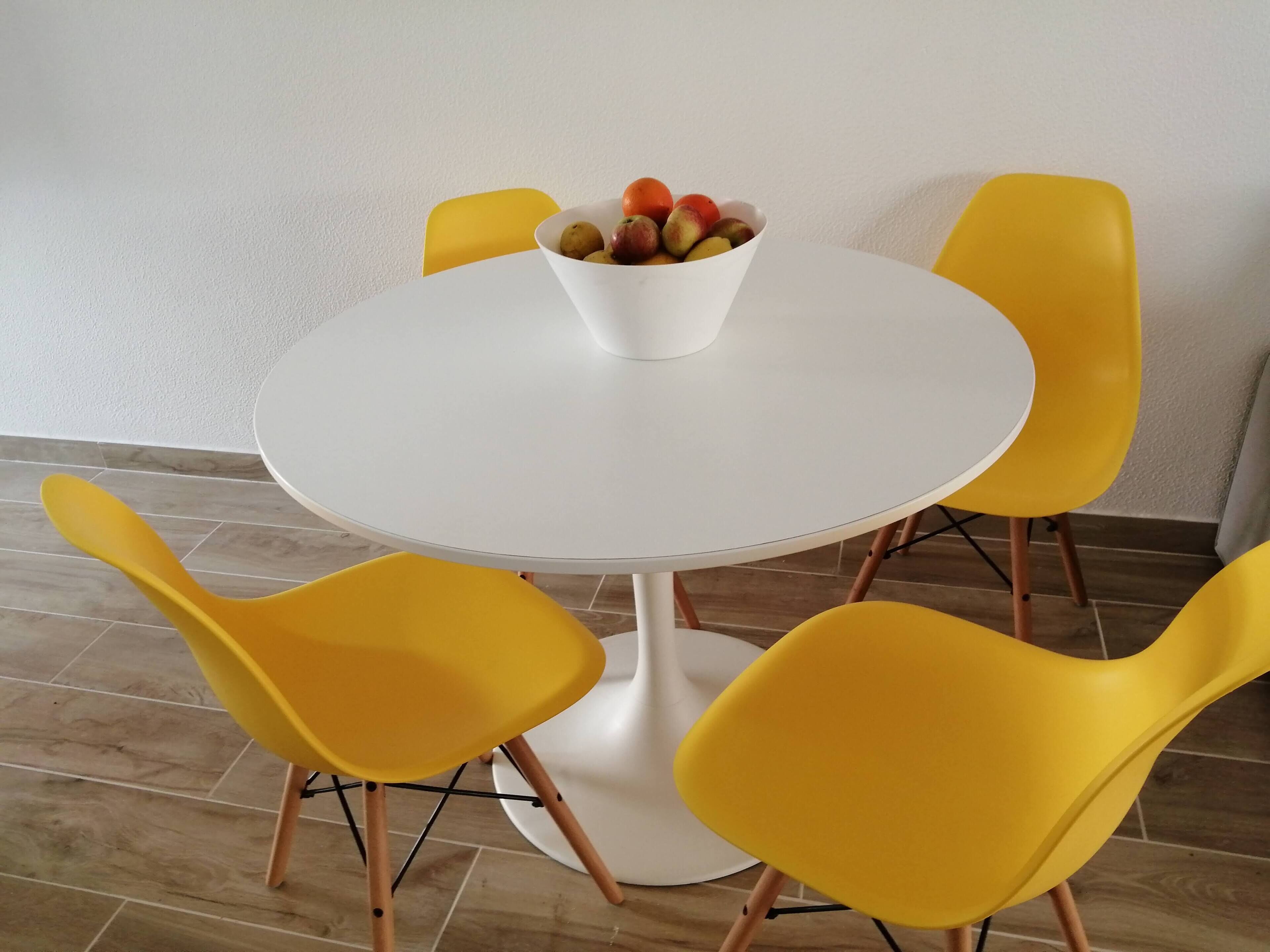 Een ronde witte tafel met een fruitschaal, omringd door vier gele stoelen.