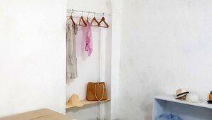 Una habitación pequeña y ordenada con una cama, un estante con un sombrero y un bolso, y un perchero con una prenda rosa.