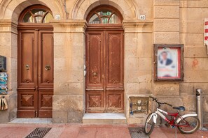 Une porte en bois à deux battants, avec un petit seuil menant à l’entrée au niveau du sol, un vélo garé à l’extérieur et un poteau de protection en métal sur la droite.
