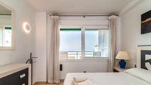 Habitación de hotel con cama, cómoda, lámpara, ventana con cortinas y vista al mar.