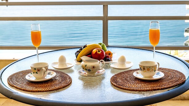 Una mesa con un frutero, dos tazas, un calentador de té y dos vasos de jugo de naranja, frente a una vista del mar.
