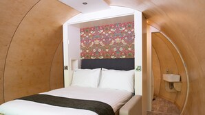Une chambre à coucher accueillante et compacte, comprenant un lit, une tête de lit ornée d’un motif floral sur le mur, une table de chevet et une petite étagère.