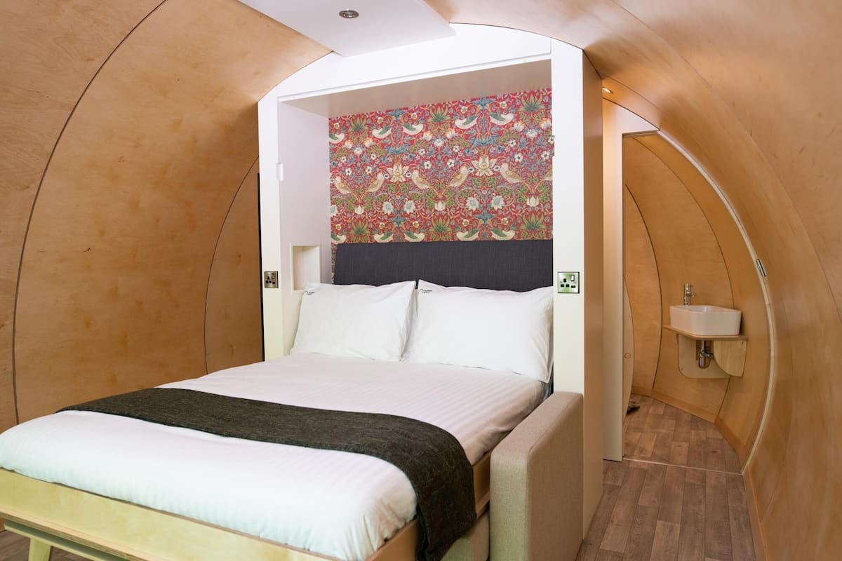 A cozy, compact bedroom with a bed, a headboard with a floral-patterned wall, a bedside table, and a small shelf.