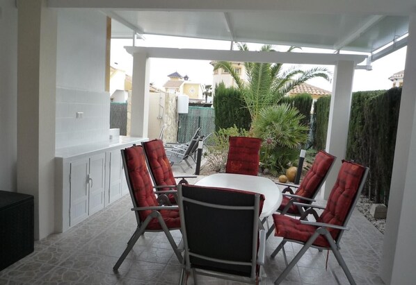 A covered patio with a round table and six red cushioned chairs, a white ceiling, and a view of a residential area with houses and greenery. - Villa 'Capricho' with Mountain View, Pool and Wi-Fi (Mazarrón)