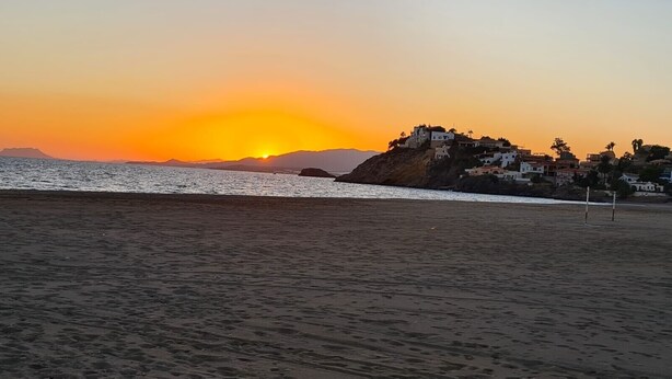 A beach with a sunset, a rocky outcrop with buildings, and calm water. - Villa 'Capricho' with Mountain View, Pool and Wi-Fi (Mazarrón)