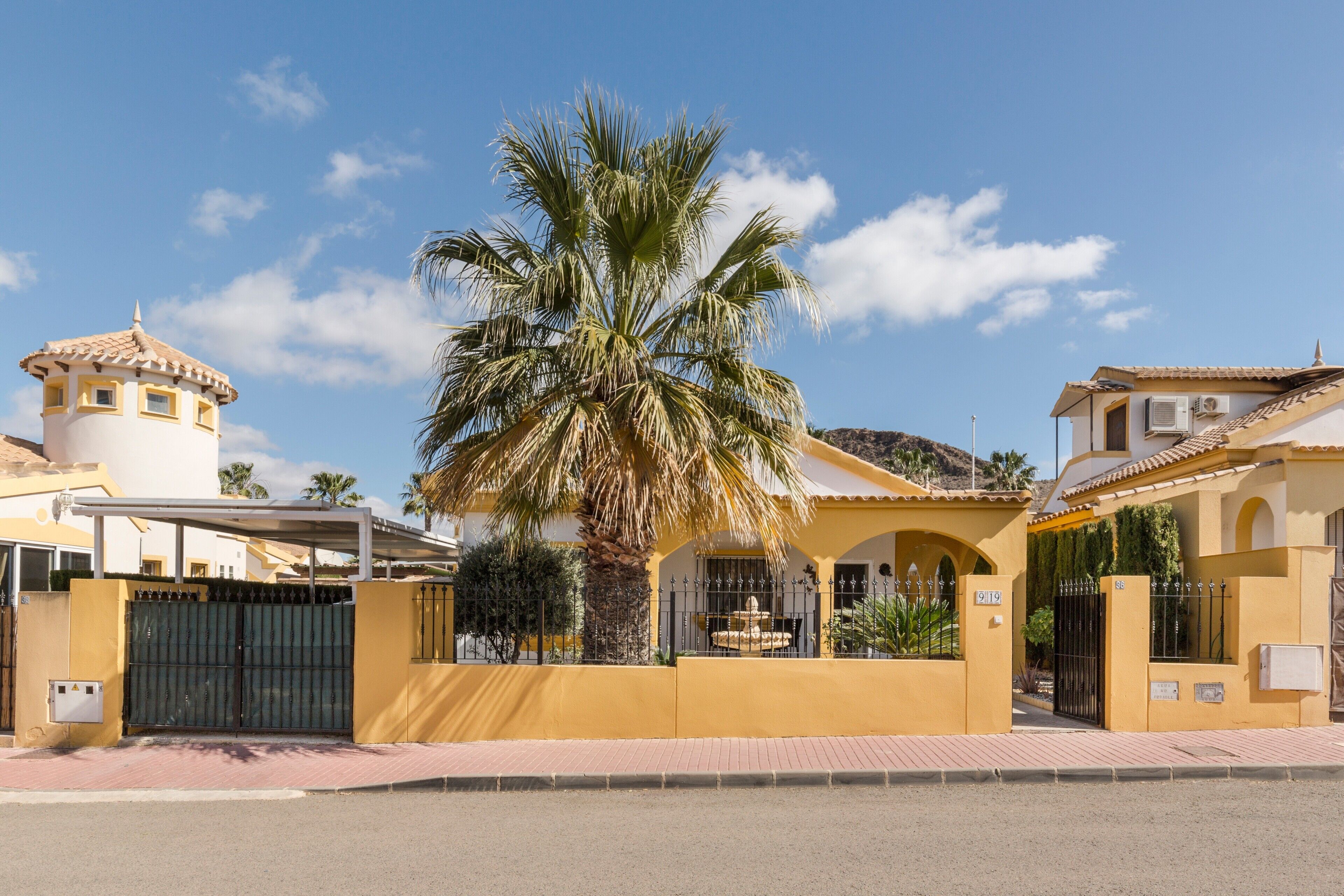 Un edificio amarillo con entrada protegida por una reja, una palmera y una estructura pequeña que recuerda a una torre.