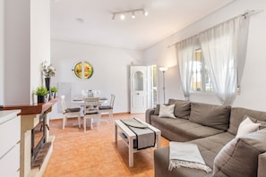A living room with a sofa, a dining table with chairs, a small table with a mat, and a door leading to another room.