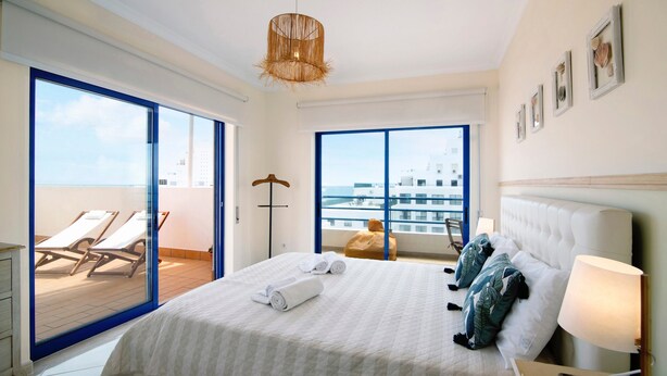 Une chambre d’hôtel avec un grand lit, une vue sur la mer, un balcon avec des chaises longues et un luminaire suspendu.