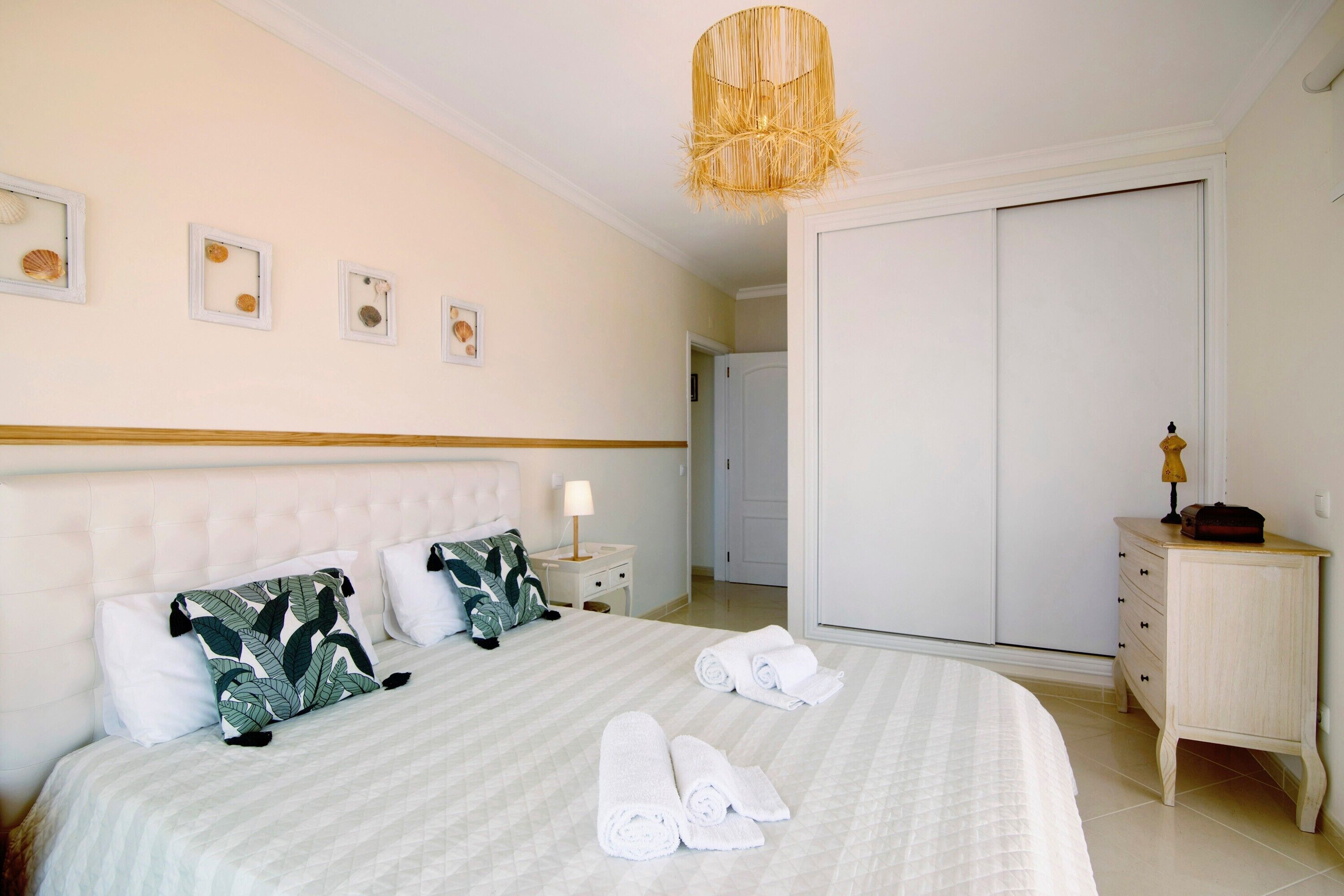 Un lit bien fait, recouvert d’une couverture à rayures, une lampe de chevet et une table de chevet avec une lampe. On peut voir des serviettes pliées sur le lit, une armoire et une petite commode en bois avec une lampe.
