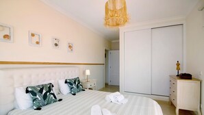 Un lit bien fait, recouvert d’une couverture à rayures, une lampe de chevet et une table de chevet avec une lampe. On peut voir des serviettes pliées sur le lit, une armoire et une petite commode en bois avec une lampe.