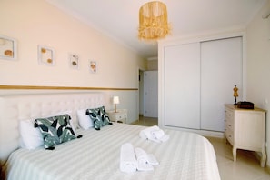 Un lit bien fait, recouvert d’une couverture à rayures, une lampe de chevet et une table de chevet avec une lampe. On peut voir des serviettes pliées sur le lit, une armoire et une petite commode en bois avec une lampe.