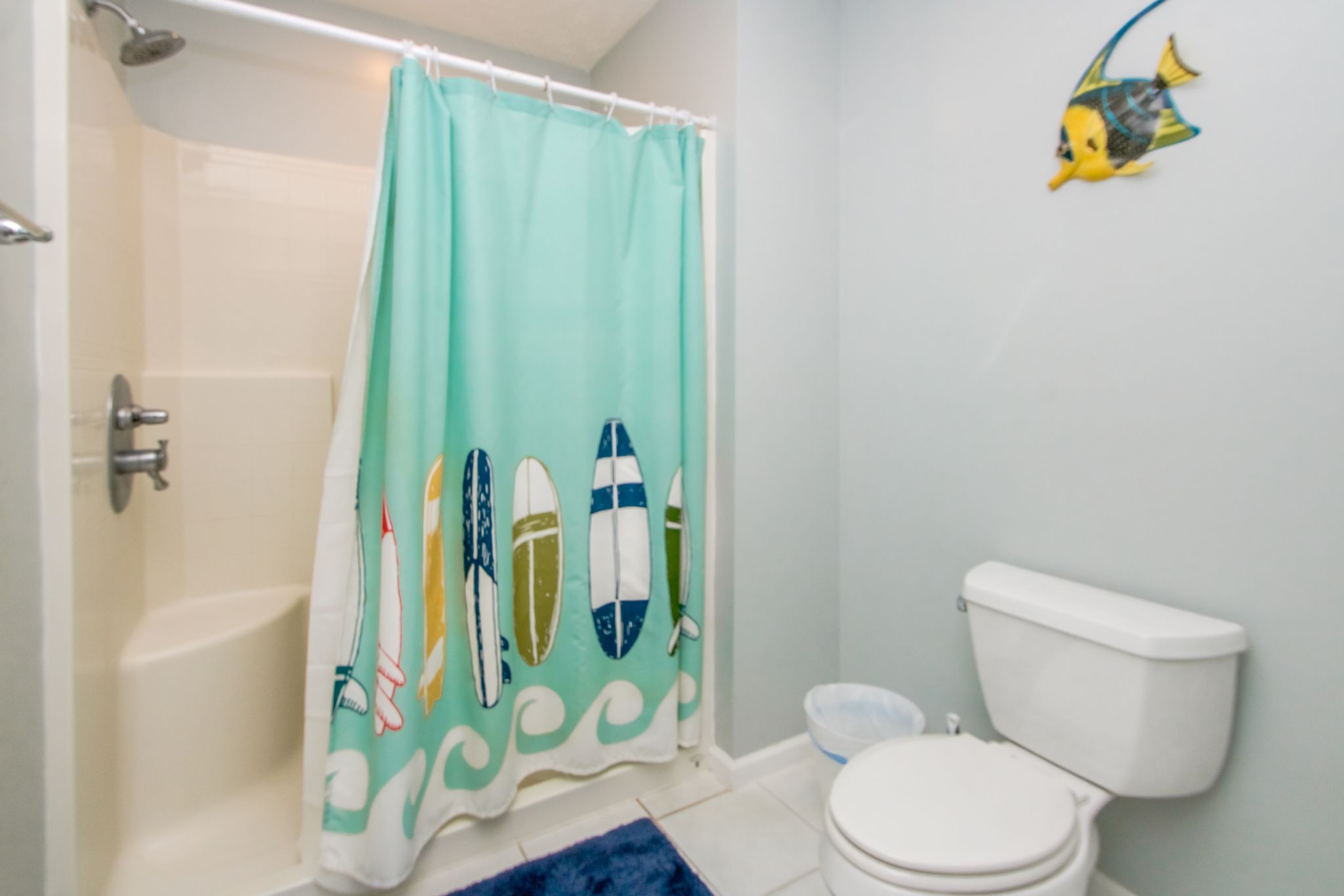 A bathroom with a shower curtain featuring surfboard designs, a toilet, and a small fish decoration on the wall.