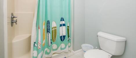 A bathroom with a shower curtain featuring surfboard designs, a toilet, and a small fish decoration on the wall.