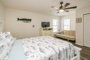 A bedroom with a bed, a TV on a stand, a ceiling fan, a window with blinds, and a door.