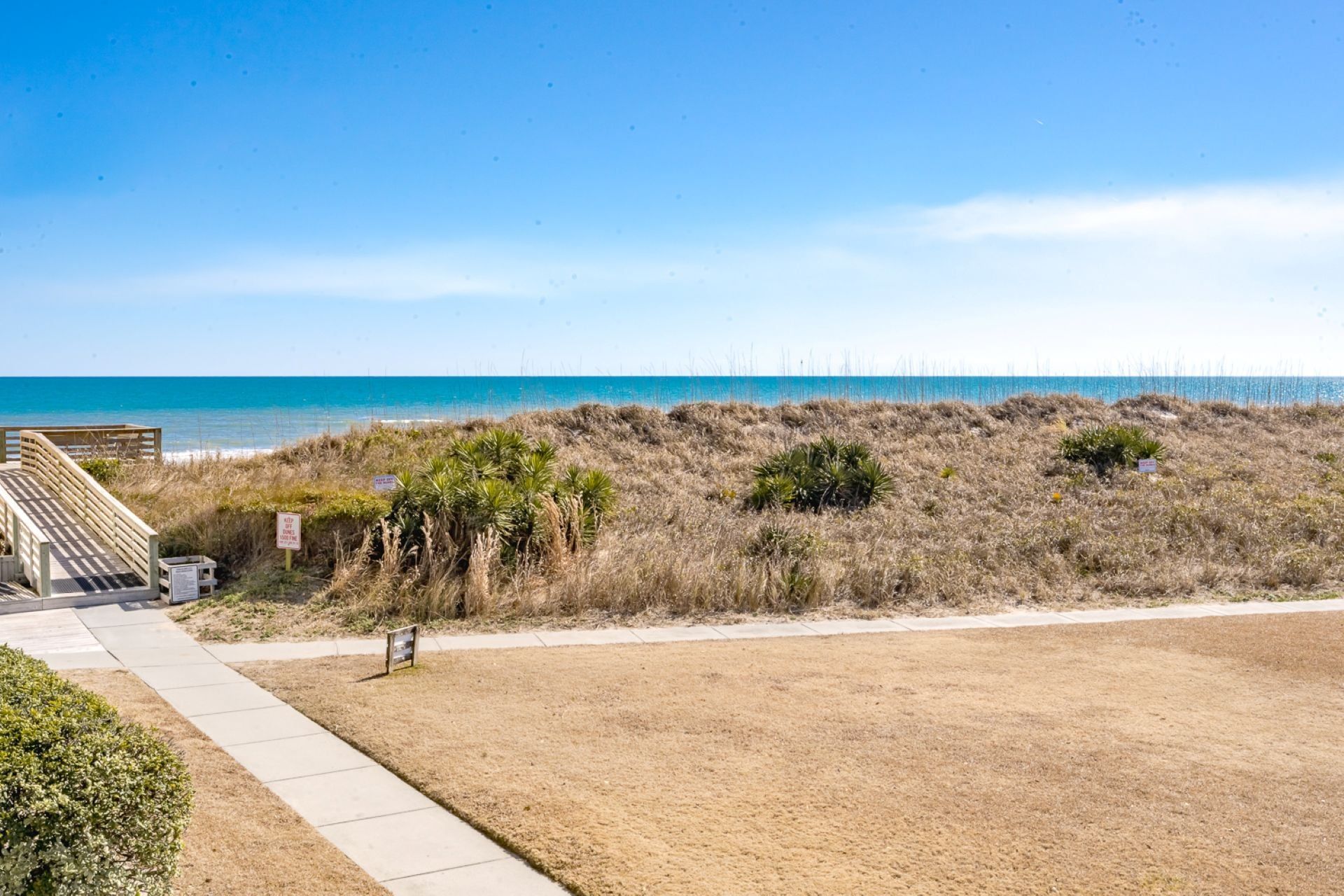 Vista de una playa con un paseo marítimo de madera que conduce al océano, un área verde con vegetación escasa y un cielo azul despejado.