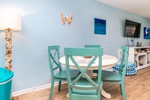 A room with teal chairs around a round table, a TV on the wall, and a bookshelf with books and decorative items. - 715 Surf - Gorgeous 1BR Surf Condo with Washer and Dryer (Surf City)