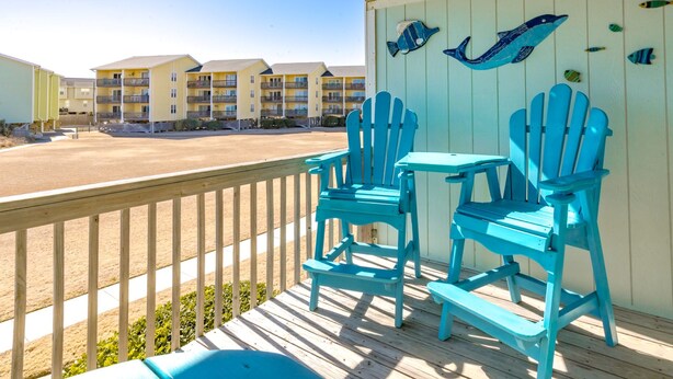 A wooden deck with two chairs and a table, overlooking a beach with buildings in the background. - 715 Surf - Gorgeous 1BR Surf Condo with Washer and Dryer (Surf City)