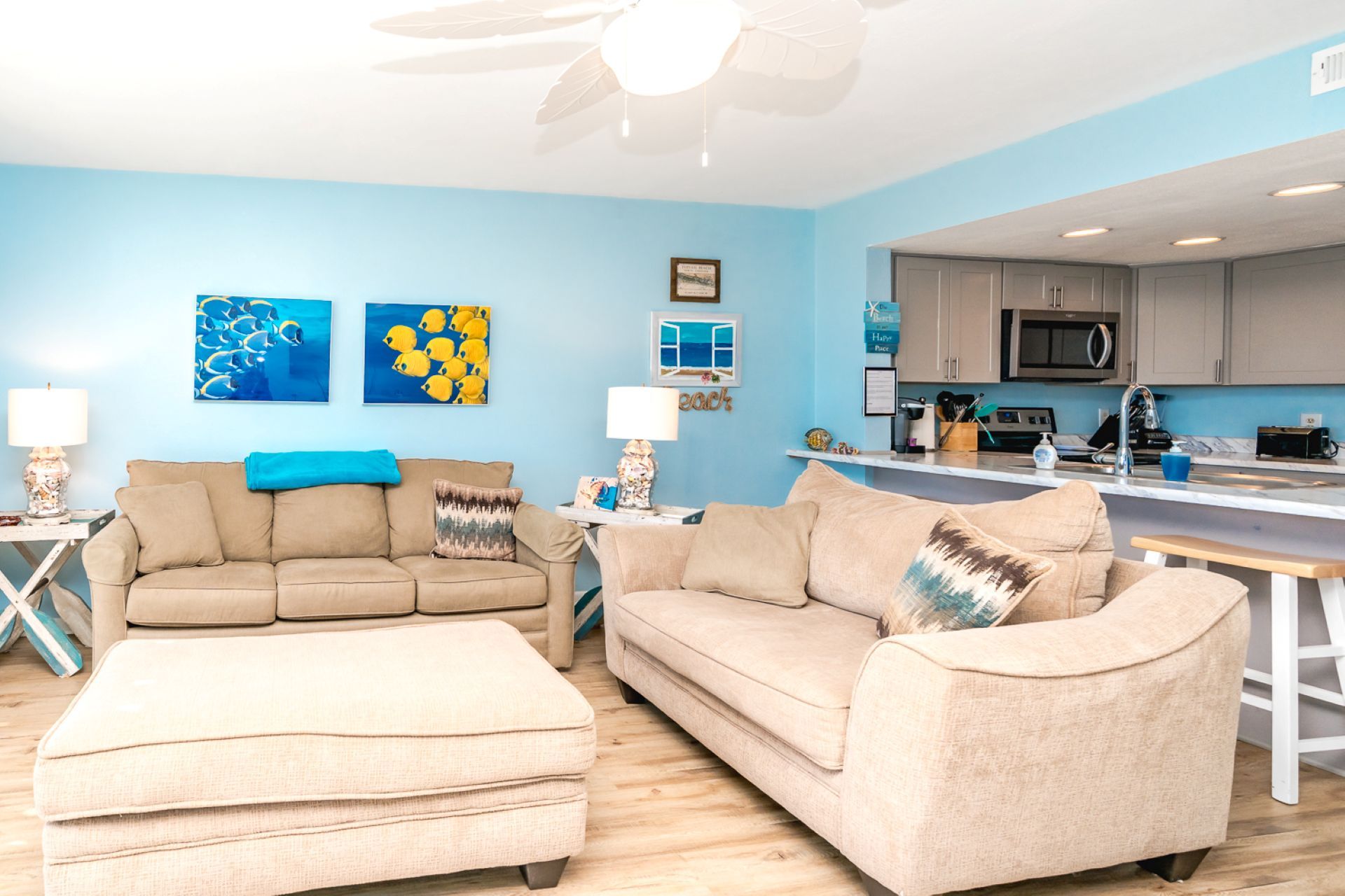 A living room with a beige sofa, a matching ottoman, and a kitchen area with a microwave and sink.