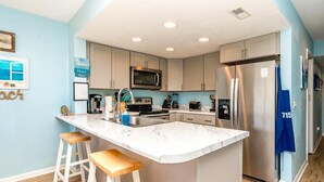 A modern kitchen with a marble island, stainless steel appliances, and wooden stools. - 715 Surf - Gorgeous 1BR Surf Condo with Washer and Dryer (Surf City)
