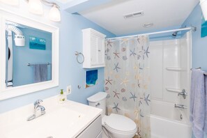 A bathroom with a white sink, a toilet, a shower with a starfish shower curtain, and a mirror with lights. - 715 Surf - Gorgeous 1BR Surf Condo with Washer and Dryer (Surf City)