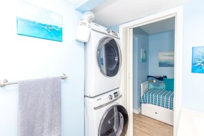 A laundry room with a stacked washer and dryer, a towel rack, and a bed with a blue and white patterned bedspread. - 715 Surf - Gorgeous 1BR Surf Condo with Washer and Dryer (Surf City)