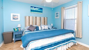 A bedroom with a bed, a nightstand, a lamp, a ceiling fan, and a window with curtains. - 715 Surf - Gorgeous 1BR Surf Condo with Washer and Dryer (Surf City)