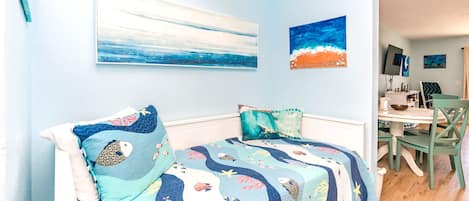 A bedroom with a bed, two pillows, a wall with two paintings, and a view into a dining area.