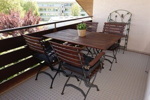 Un balcon avec une table et des chaises en bois, une plante en pot et une chaise en métal ornée d’un motif décoratif.