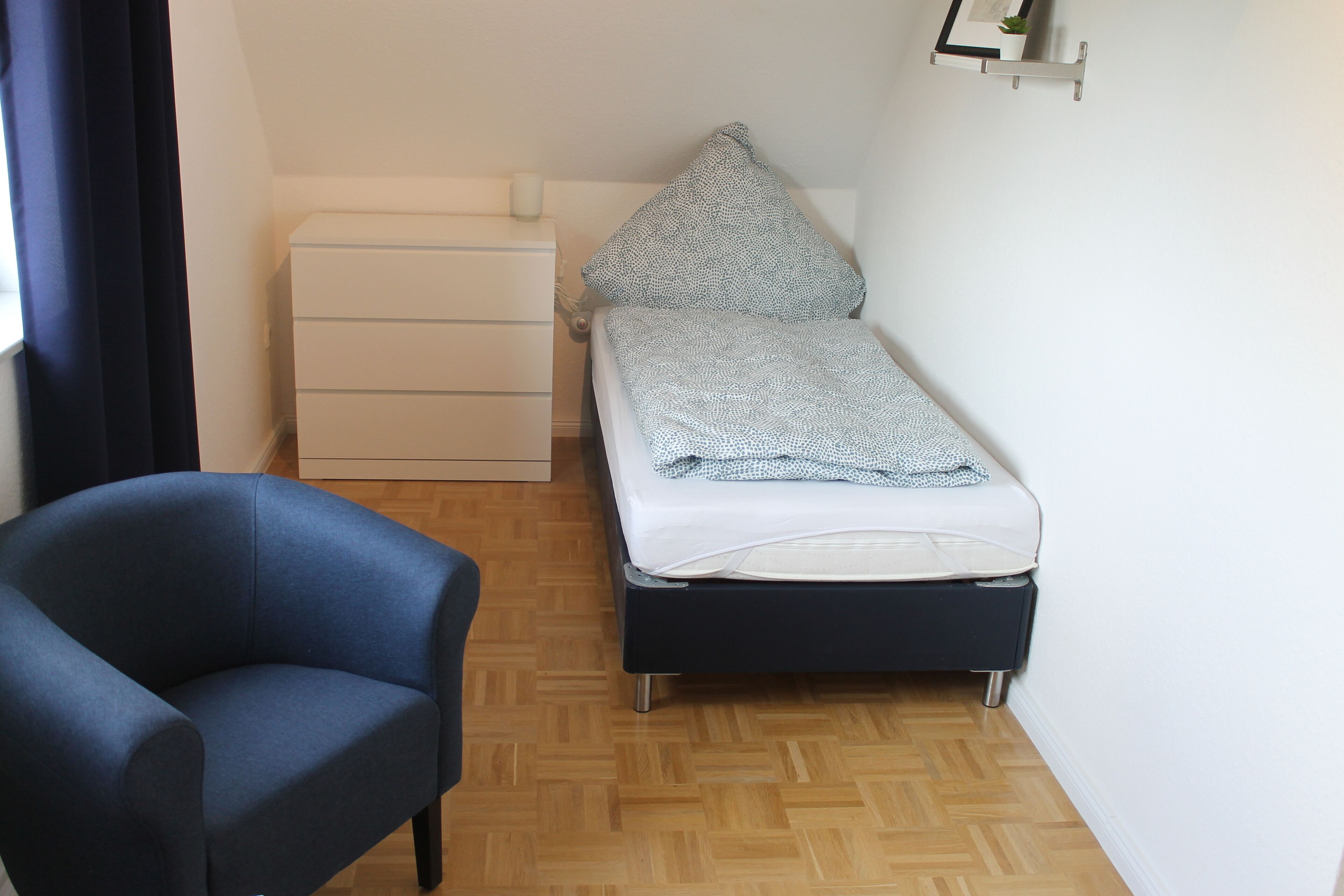 A small, neatly arranged room with a single bed, a chair, a bedside table, and a wall-mounted shelf.