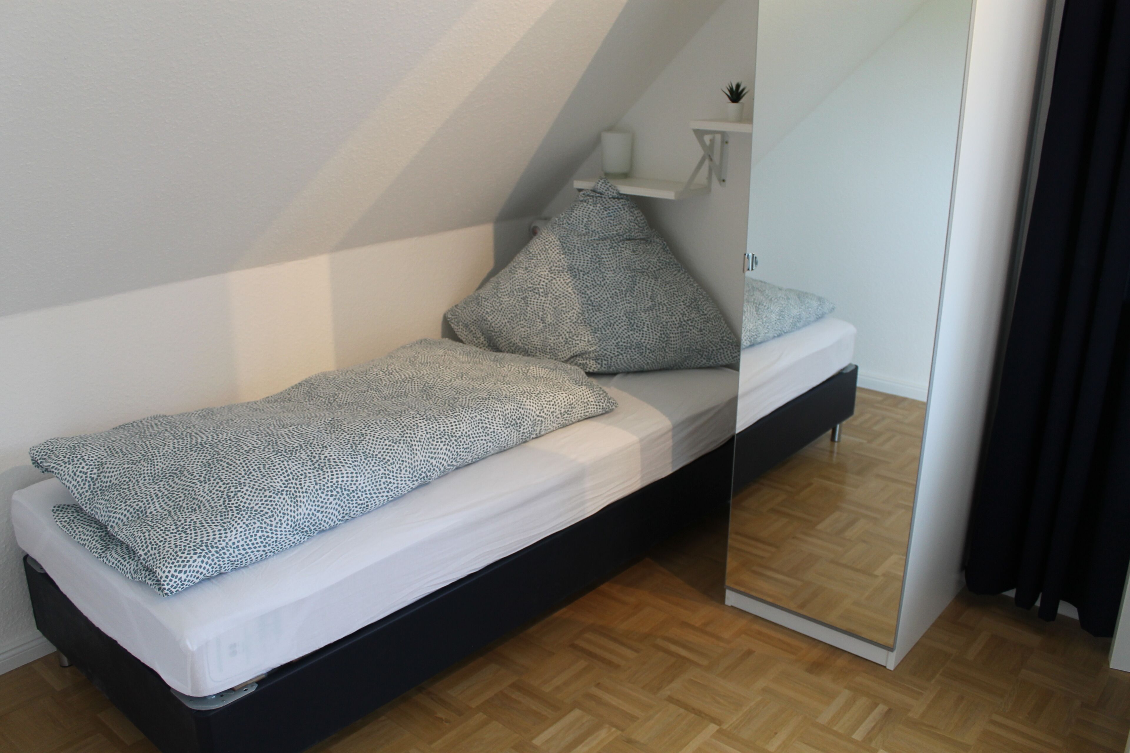 A small, modern room with a bed, a bedside table, a mirror, and a small shelf with a plant.