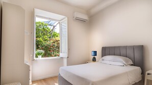 Ein Schlafzimmer mit einem Bett, einem Nachttisch, einer Lampe, einer Klimaanlage und einem Fenster mit Blick ins Grüne.