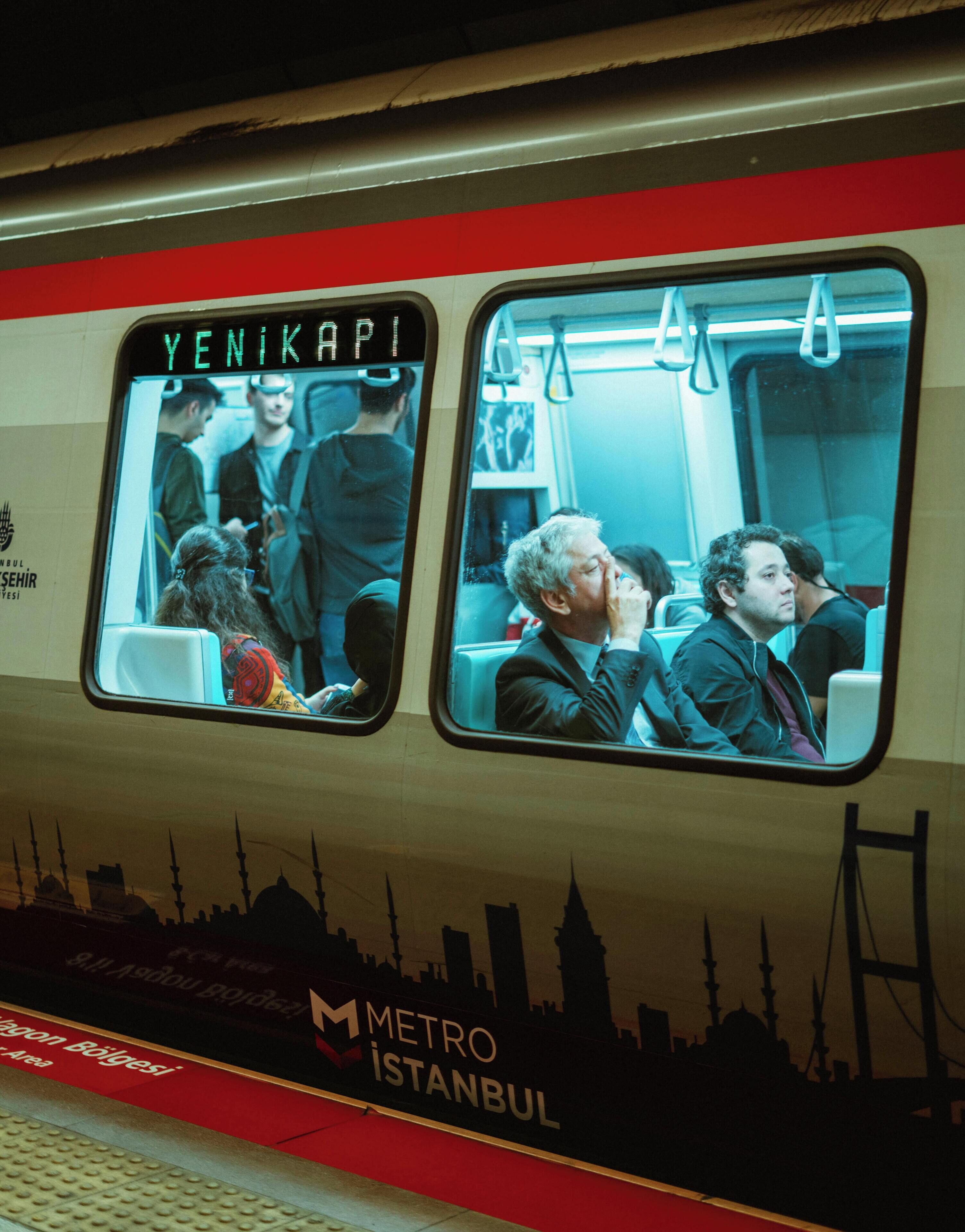 Een treininterieur met passagiers, zittend en staand, een raam met uitzicht op een stadsgezicht, en een bord waarop 