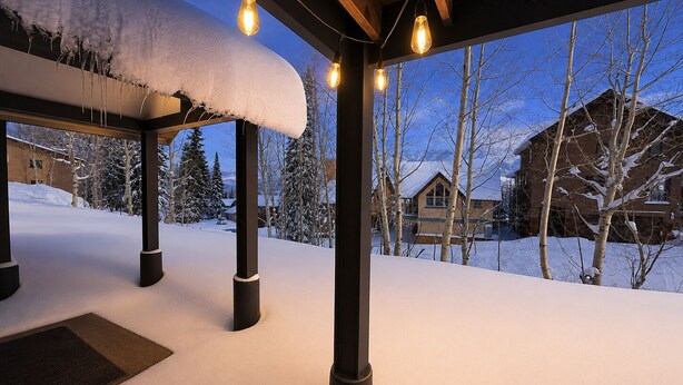 Un espace extérieur enneigé, agrémenté de poutres en bois et de guirlandes lumineuses, une structure en bois et une vue sur des maisons et des arbres recouverts de neige.