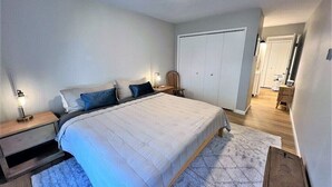 Une chambre à coucher avec un lit, des tables de chevet, une chaise, une armoire et une salle de bain visible au fond.