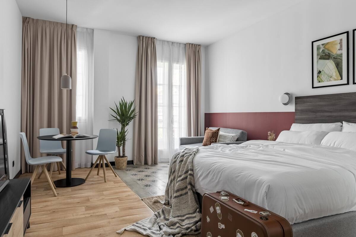 Habitación de hotel moderna con una cama grande, un escritorio con sillas, una planta en maceta y vistas a la ciudad.