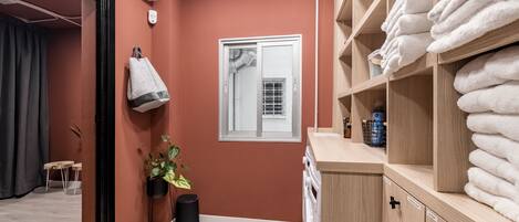A hotel room with a red wall, a window with a view, a towel rack, a plant, and a wooden cabinet with towels.