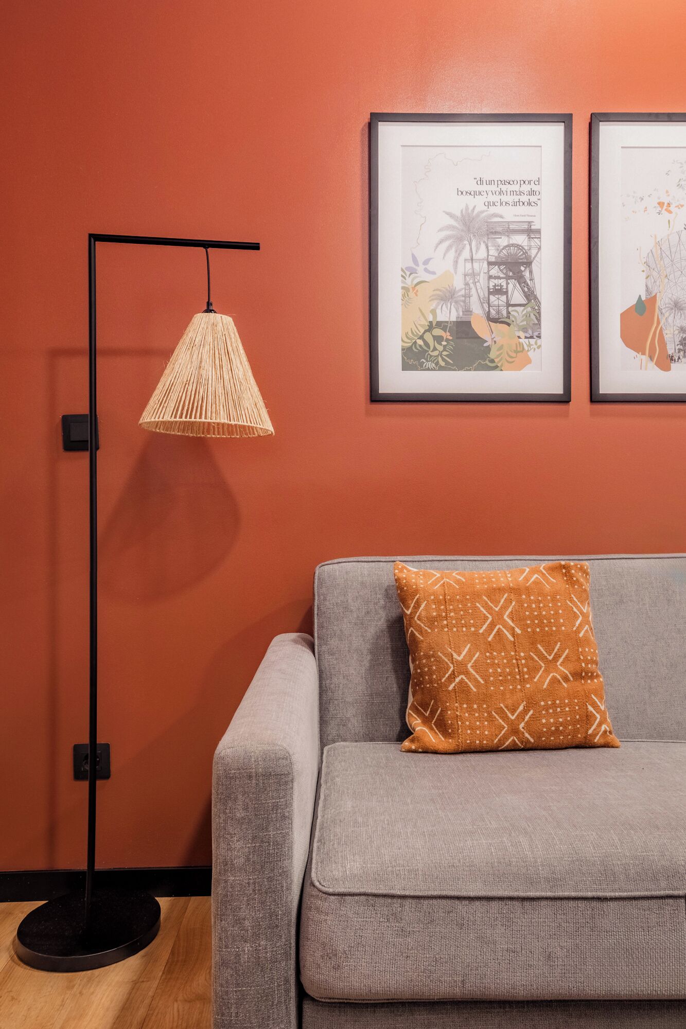 Ein Zimmer mit grauem Sofa, einem orangefarbenen Kissen, einer Stehlampe und zwei gerahmten Bildern an der Wand.