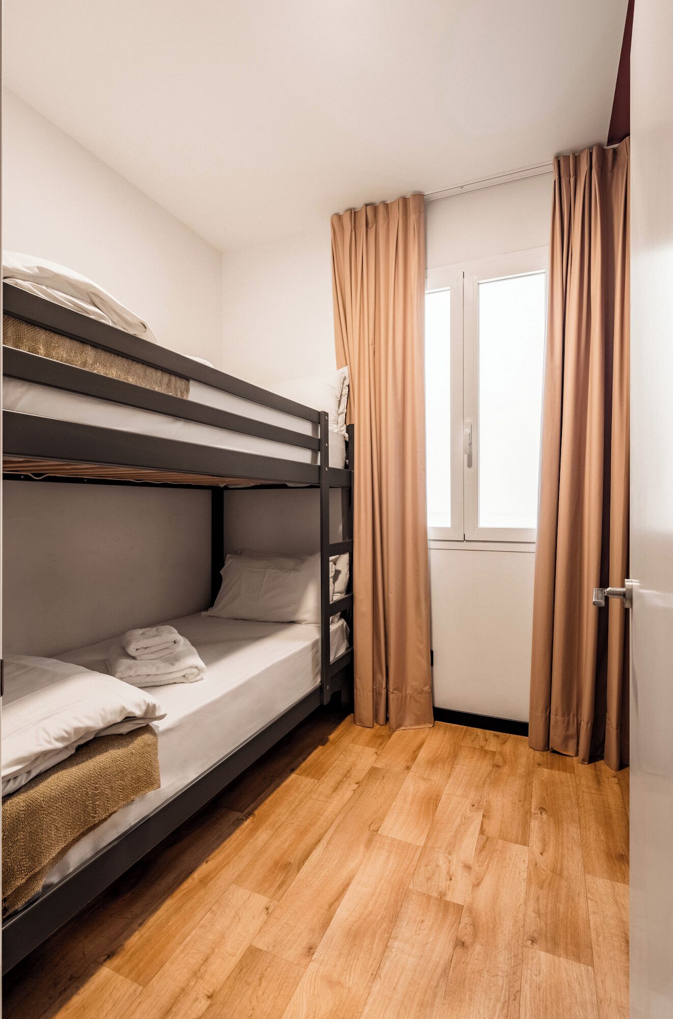 A bunk bed room with a wooden floor, a window with curtains, and a door.