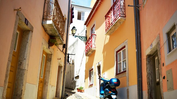 A narrow street with buildings on either side, a scooter parked on the right, and a street lamp in the center.