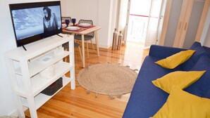 A living room with a television, a blue sofa with yellow cushions, a woven rug, and a dining table with chairs.