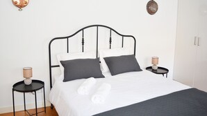 A neatly made bed with white and dark gray linens, two bedside tables with lamps, and a white wardrobe in a minimalist room.