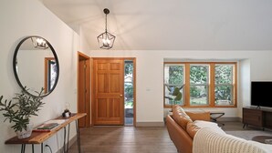 Ein großzügiger Wohnbereich mit Holzboden, einem großen Spiegel, einer Holztür, einem Fenster mit Jalousien, einem Ledersofa und einem Fernseher auf einer Holzkommode.