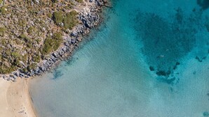 Vue aérienne d’une plage aux eaux turquoise et limpides, avec un terrain rocheux et une rangée de parasols bleus sur le sable.