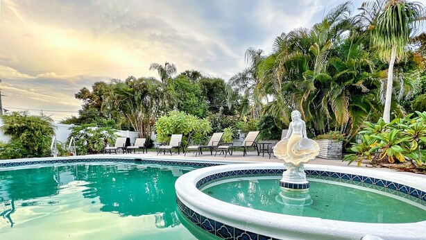 A swimming pool with a decorative fountain, surrounded by lounge chairs and tropical plants. - Fun Getaway in the Sun Private Saltwater Pool Yard (Gulfport)