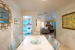 A dining area with a long table, chairs, and a floral table runner. There is a kitchen in the background with blue cabinets and a microwave.