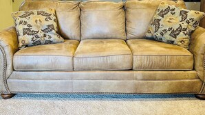 A beige leather sofa with floral-patterned cushions in a living room with a bookshelf and kitchen in the background.