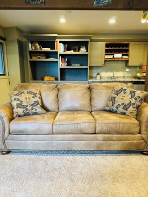A beige leather sofa with floral-patterned cushions in a living room with a bookshelf and kitchen in the background.