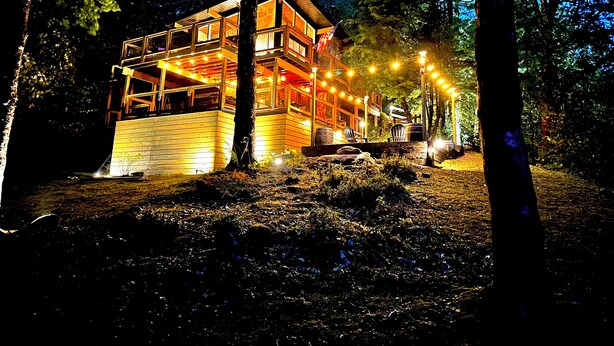 A wooden cabin with multiple levels, surrounded by trees and lit by string lights.