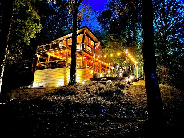 A wooden cabin with multiple levels, surrounded by trees and lit by string lights.
