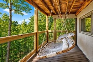 Una terraza de madera con una silla hamaca colgante, baranda de madera y vistas a un bosque.